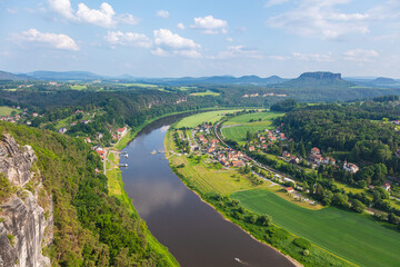 Scenic panoramic view from the Bastei cliffs over the Elbe River valley with sandstone rocks, lush forests, rural village rooftops, distant mountain ranges under a vibrant summer sky. Saxony, Germany