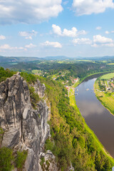 Scenic panoramic view from the Bastei cliffs over the Elbe River valley with sandstone rocks, lush forests, rural village rooftops, distant mountain ranges under a vibrant summer sky. Saxony, Germany