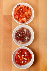 Three bowls arranged vertically on a wooden surface, each containing a different salad: roasted pepper, tomato, and garlic salad at the top, bean salad in the middle, and roasted pepper and garlic