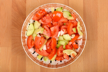 A bowl of freshly chopped tomato, cucumber, bell pepper, and onion salad on a wooden background, photographed from a top-down angle