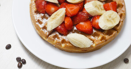 Crispy golden waffle crowned with sliced strawberries and bananas, dusted with powdered sugar, resting on white plate near scattered coffee beans, morning indulgence