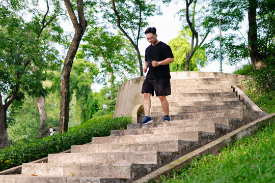Young Asian man walks down outdoor stairs in a lush green park on a sunny day amidst trees and people enjoying the fresh air