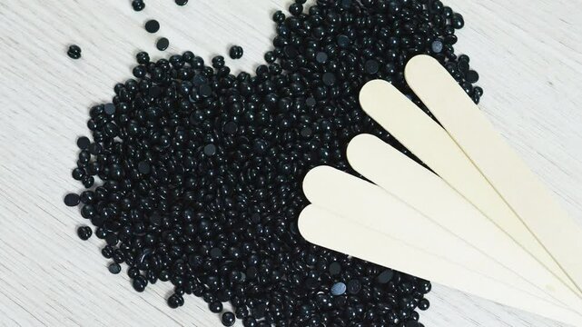 Black hard wax beans forming heart pattern on rustic wooden surface, positioned near wooden spatulas, highlighting hair removal beauty treatment setup