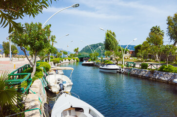 Scenic canal with moored boats lined by decorative street lights and lush greenery in Icmeler, Marmaris, Mugla Province, Turkey. Bright sunny day with clear blue sky and distant mountains.