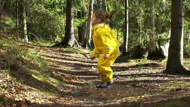 Happy child in yellow rain suit jumping in puddle in park on a sunny day. Active outdoor play, fun and nature exploration in Scandinavian woodland environment.