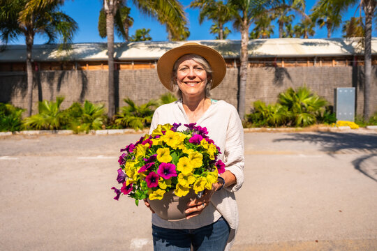 Senior woman holding colorful petunias in a pot, smiling at the botanical garden