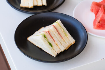 Close-up of a sandwich with ham, cucumber, and white bread on a black plate with watermelon slices...