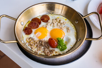Close-up of a skillet breakfast with fried eggs, sausage, tomatoes, and herbs served in a metal pan on a white tabletop