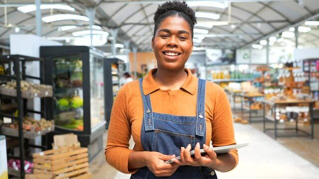 Confident young african american saleswoman wearing denim overalls and holding digital tablet while standing in grocery store and looking at camera