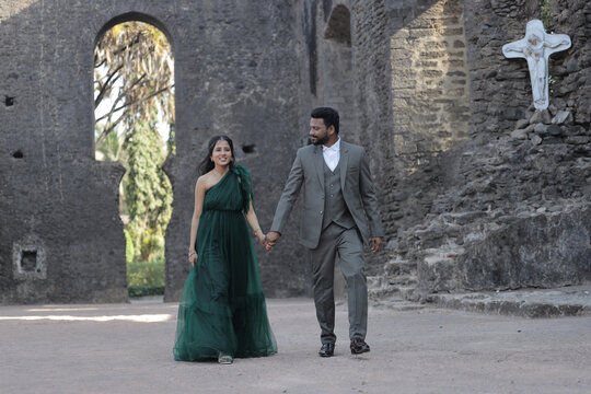 A stylish Indian couple poses affectionately at an ancient stone monument. The woman wears a flowing dark green gown, and the man is dressed in a classic three-piece suit.