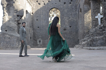 Naklejka premium A stylish Indian couple poses affectionately at an ancient stone monument. The woman wears a flowing dark green gown, and the man is dressed in a classic three-piece suit.