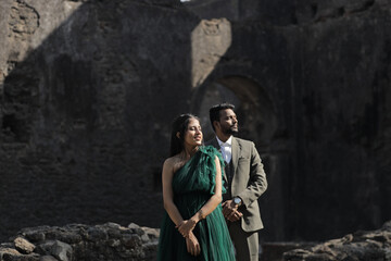 A stylish Indian couple poses affectionately at an ancient stone monument. The woman wears a flowing dark green gown, and the man is dressed in a classic three-piece suit.