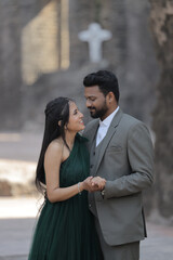 A stylish Indian couple poses affectionately at an ancient stone monument. The woman wears a flowing dark green gown, and the man is dressed in a classic three-piece suit.