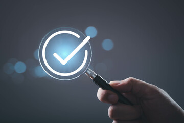 A hand holding a magnifying glass with a checkmark icon, symbolizing verification or confirmation...