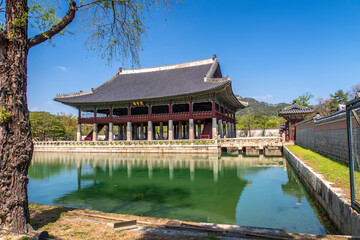 A traditional Korean pavilion, Gyeonghoeru, standing on stilts over a tranquil green pond with reflections, under a clear blue sky.
