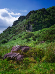 Strickeen mountain face covered in lush greenery and rocks on a bright, partly cloudy day in County Kerry, Ireland.
