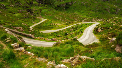 A narrow road winds sharply through a verdant, hilly landscape. Rocks and tall grass grow alongside the road. The Gap of Dunloe shows its beautiful scenery.