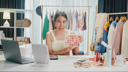 Young beautiful Asia lady blogger looking at camera recording swatch apply eyeshadow palette on skin show review to follower product vlog video live streaming at home. Business online media concept.