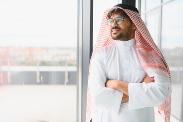 Confident arab businessman wearing traditional kandura and looking out window in modern office