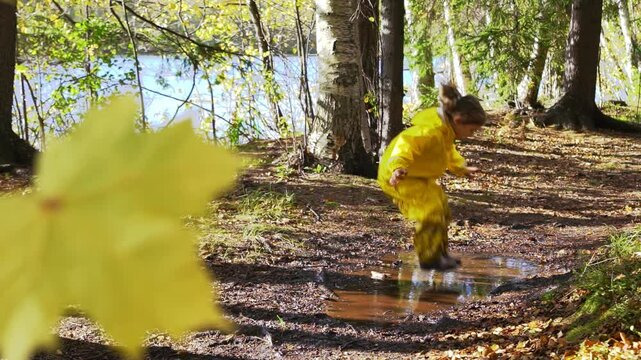 Happy child in yellow rain suit jumping in puddle in park on a sunny day. Active outdoor play, fun and nature exploration in Scandinavian woodland environment.