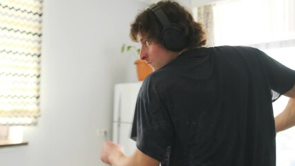 Young guy dancing alone at home in wireless headphones at home kitchen. Close-up shot shows rhythmic arm movements and emotional expression. Casual clothing, cozy background.