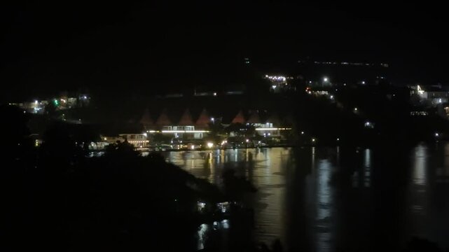 Nighttime view of traditional Batak-style buildings illuminated along the shore of Lake Toba, North Sumatra, with shimmering reflections on the calm water and glowing city lights.

