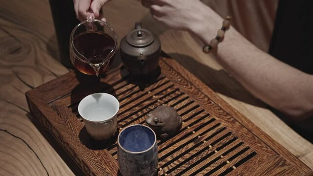 A tea master carefully pours brewed tea from a clay teapot into a glass teapot during a traditional ceremony on a wooden tray.