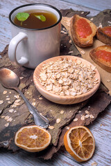 Rustic healthy breakfast with rolled oats, herbal tea in a white mug, toasted bread with tomato, and dried orange slices on a wooden bark surface