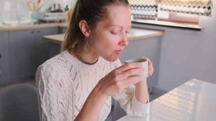 Woman in a cozy white sweater drinking a hot beverage from a white cup, holding it with both hands in a bright kitchen. Enjoying a warm drink at home. Close-up. - Powered by Adobe