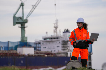woman marine engineers wearing safety gear working on a dockside project using a laptop, with a large cargo ship and industrial crane in the background under cloudy sky.