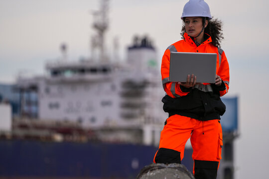woman marine engineers wearing safety gear working on a dockside project using a laptop, with a large cargo ship and industrial crane in the background under cloudy sky. - Powered by Adobe
