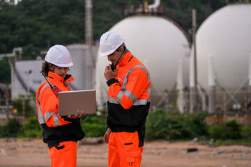 Two industrial engineers in safety suits working at a coastal energy facility. They discuss inspection data with a laptop and walkie talkie near large spherical gas storage tanks.