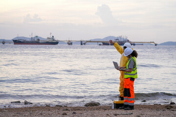 Two environmental engineers perform ocean water sampling and digital analysis near an industrial port. One holds a test tube while the other enters data on a laptop by the shoreline.