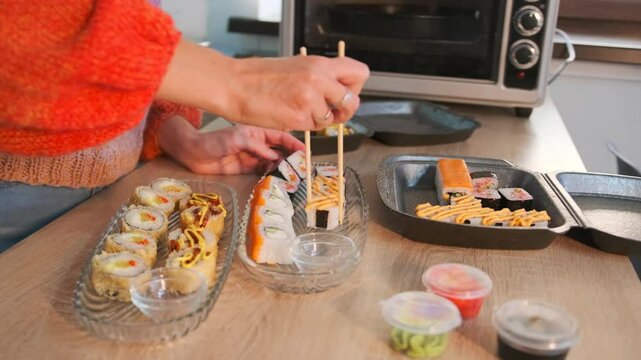 Female hands in a bright sweater take delicious sushi rolls from a delivery box with chopsticks serving transparent plates in a modern kitchen, takeaway food from japan restaurant. - Powered by Adobe