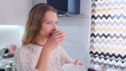 A tight shot of a beautiful woman gently cradling a white ceramic cup, bringing it to her lips to take a sip of a dark drink, coffee or tea, near the window.