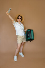 Young girl traveler in summer outfit and hat with suitcase in hands, ready for vacation. Standing on beige background with space for text.