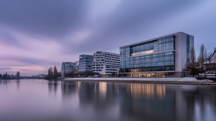 Tranquil urban waterfront at dusk with modern illuminated buildings, calm reflective water, pastel-hued sky, snowy ground, leafless trees, and distant bridge.