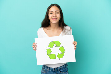 Young French woman isolated on blue background holding a placard with recycle icon with happy...