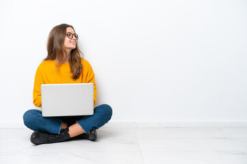 Young caucasian woman with a laptop sitting on the floor isolated on white background looking side