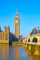 Fototapeta na wymiar Big Ben stands tall against a clear blue sky, reflecting sunlight on the River Thames. The iconic clock tower is surrounded by historic buildings, marking London's rich heritage.
