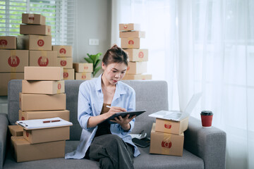 An Asian woman uses a tablet to manage online orders and prepare packages for shipping —...