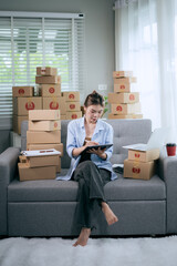 An Asian woman uses a tablet to manage online orders and prepare packages for shipping — reflecting the growth of SME businesses and the rising trend of online shopping.