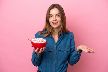 Young caucasian woman holding a bowl of cereals isolated on pink background with shocked facial...