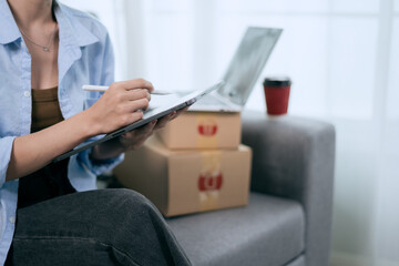 An Asian woman uses a tablet to manage online orders and prepare packages for shipping —...