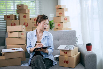 An Asian woman uses a tablet to manage online orders and prepare packages for shipping —...