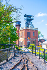 At Sevcinsky mine in Brezove hory, Pribram, Czechia, visitors explore a historical site with striking architecture and mining equipment surrounded by greenery and a clear blue sky.