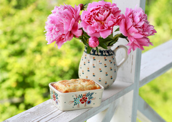 Cottage cheese casserole in a beautiful ceramic baking dish on the railing of the white terrace, outside. In the background is a jug with pink peonies. Selective focus.