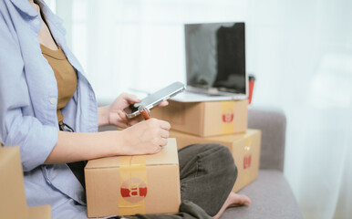 An Asian female entrepreneur prepares parcels and checks online orders from her home using her laptop and smartphone, reflecting the concept of online shopping and starting a SME business.