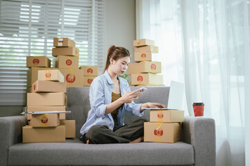 An Asian woman entrepreneur prepares parcels and manages her product orders via smartphone, ready to ship them to customers — reflecting the rise of online shopping and small business startups.