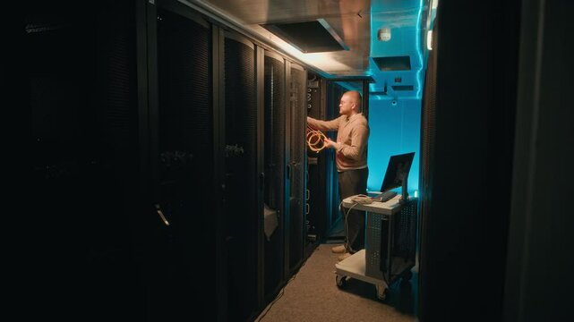 Very long shot of male Caucasian specialist checking cables in supercomputing equipment while working in server room